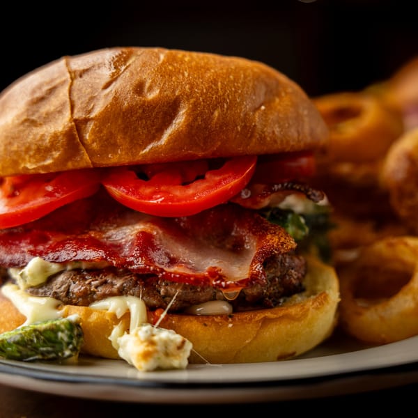Close-up of a Sterling's bacon cheeseburger with tomatoes, melted cheese, and a side of onion rings.