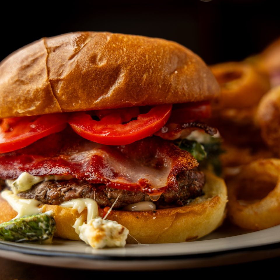 Close-up of a Sterling's bacon cheeseburger with tomatoes, melted cheese, and a side of onion rings.