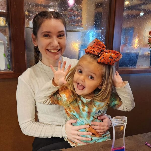 A smiling woman holds a playful child with an orange bow, both looking at the camera.