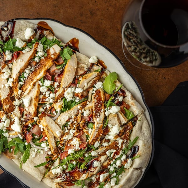 Overhead view of a flatbread pizza topped with chicken, arugula, feta, and balsamic glaze.