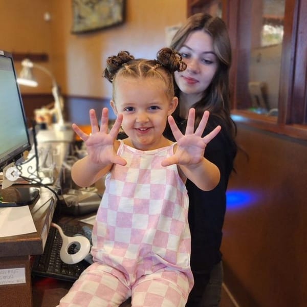 A young girl in a pink and white checkered outfit smiles, holding up her hands, with a teen behind her.