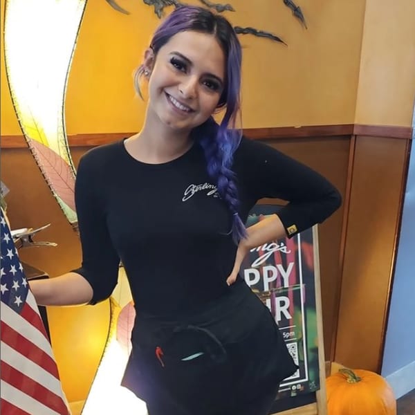 A smiling Sterling's Restaurant server with purple hair stands next to an American flag and a pumpkin.