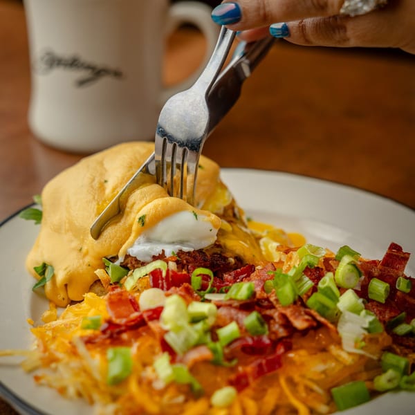 A fork and knife cutting into Sterling's breakfast dish with eggs, hash browns, bacon, and green onions.