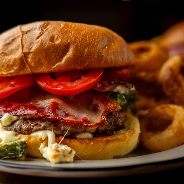 A close-up of a Sterling's burger with bacon, tomatoes, and melted cheese, served with onion rings.