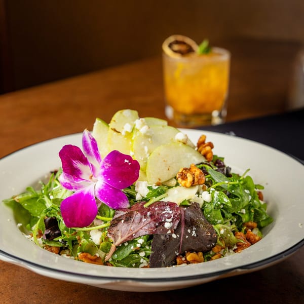 A vibrant salad with greens, sliced apples, walnuts, feta, and a purple orchid garnish.