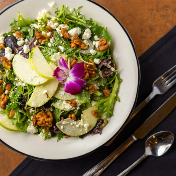 A vibrant salad with mixed greens, apple slices, walnuts, feta cheese, and an orchid garnish.