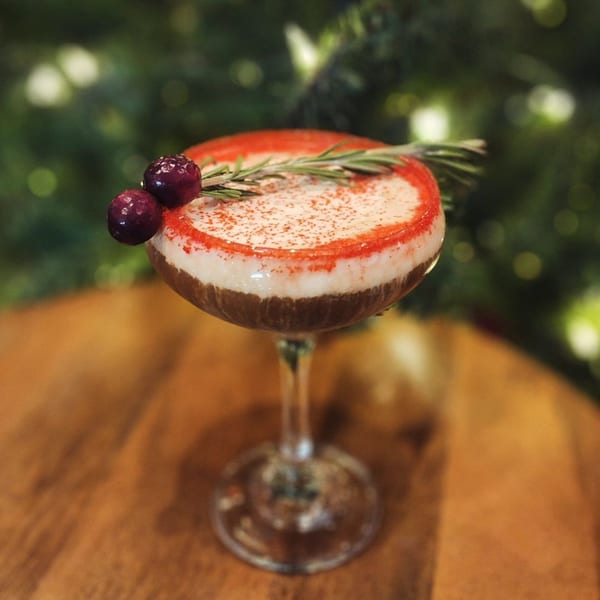 A festive cocktail with red sugar rim, white foam, cranberries, and rosemary sprig.