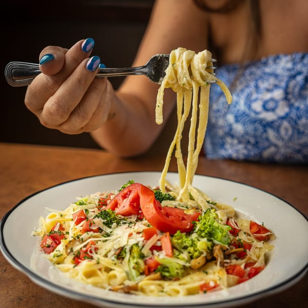 A hand with blue nail polish lifts creamy pasta with a fork from a plate of pasta, tomatoes, and broccoli.