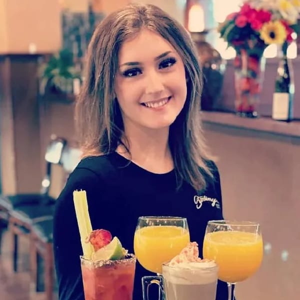 Smiling Sterling's Restaurant server holding a tray with breakfast drinks: Bloody Mary, orange juice, and hot chocolate.