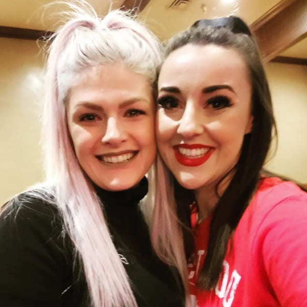 Two smiling women, one with light pink hair and one with dark hair and red lipstick, pose together.