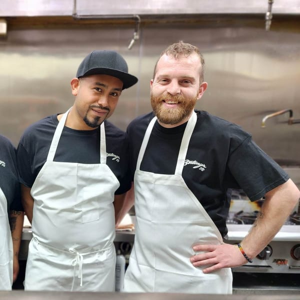 Two smiling Sterling's Restaurant chefs in black shirts and white aprons in a commercial kitchen.